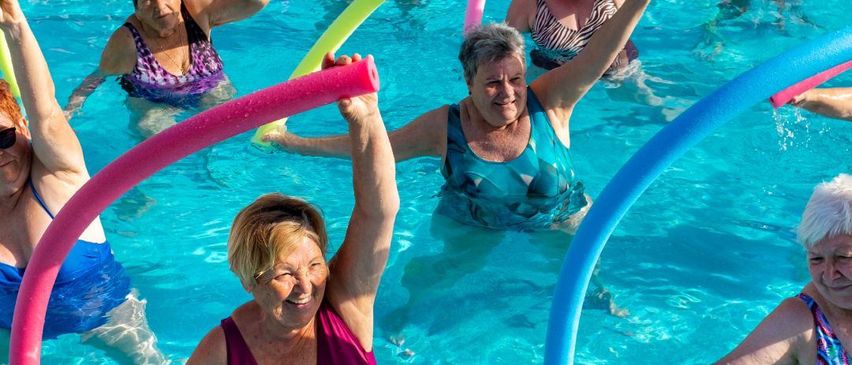 Eine Gruppe von Frauen schwimmt in einem Pool mit bunten Übungsgeräten. Sie lächeln und scheinen es zu genießen.