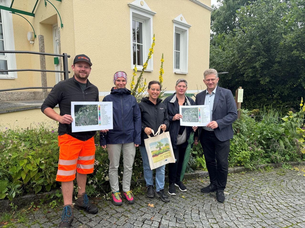 Fünf Personen stehen vor einem Haus mit gelber Außenwand und weißen Fenstern. Sie halten Karten und eine Tasche und lächeln für ein Foto.