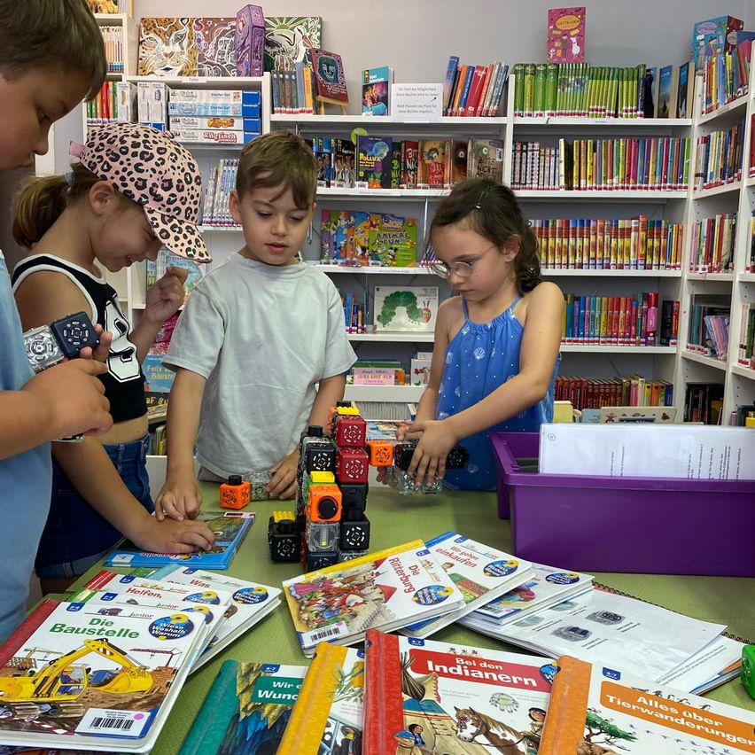Vier Kinder spielen mit Würfeln und Büchern auf einem Tisch in einer Bibliothek. Sie sind in einer Aktivität verwickelt, möglicherweise ein Spiel oder Lernen. Die Kinder tragen Freizeitkleidung, und die Bibliothek ist gefüllt mit bunten Büchern an Regalen.