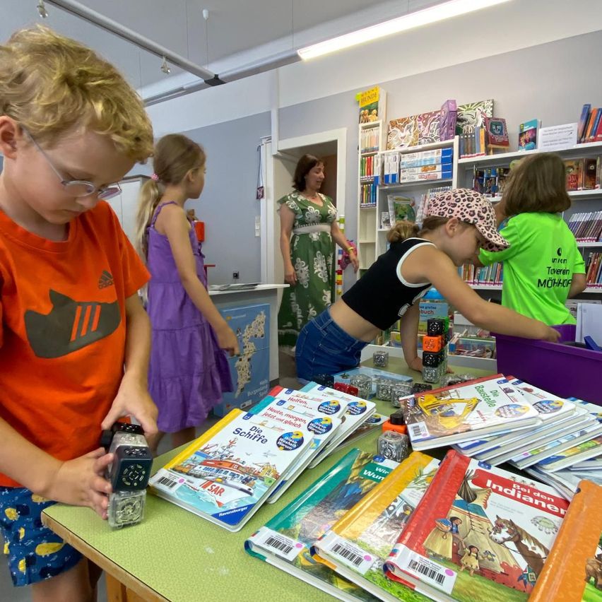 Kinder versammeln sich um einen Tisch voller Bücher in einer Bibliothek. Ein Junge untersucht eine kleine Schachtel, während ein Mädchen sich beugt, um ein Buch anzuschauen. Hinter ihnen steht eine Frau in der Nähe einer Tür.