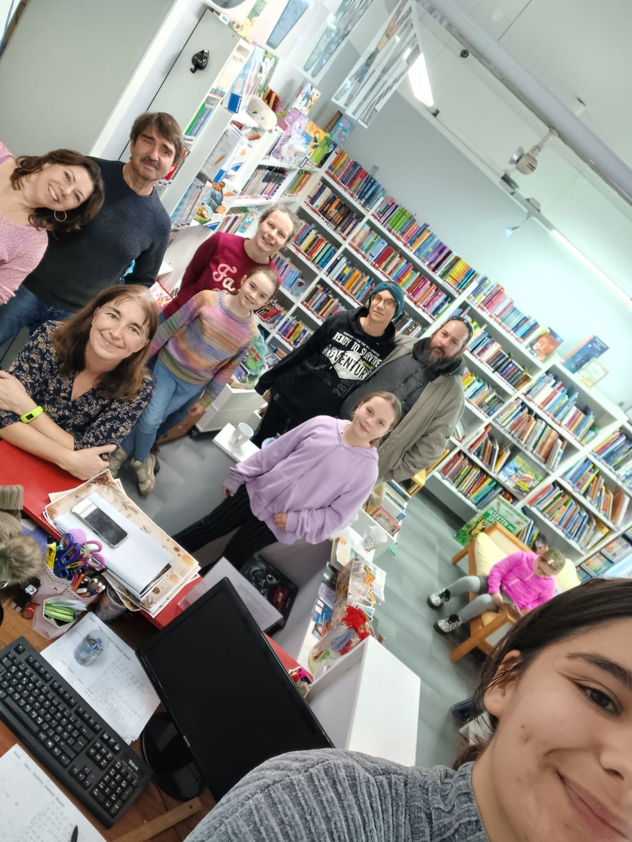 Eine Gruppe von Menschen in einer Bibliothek, die lächelnd für ein Foto posieren. Regale voller Bücher umgeben sie. Ein Kind sitzt auf einem Stuhl in der Ecke.