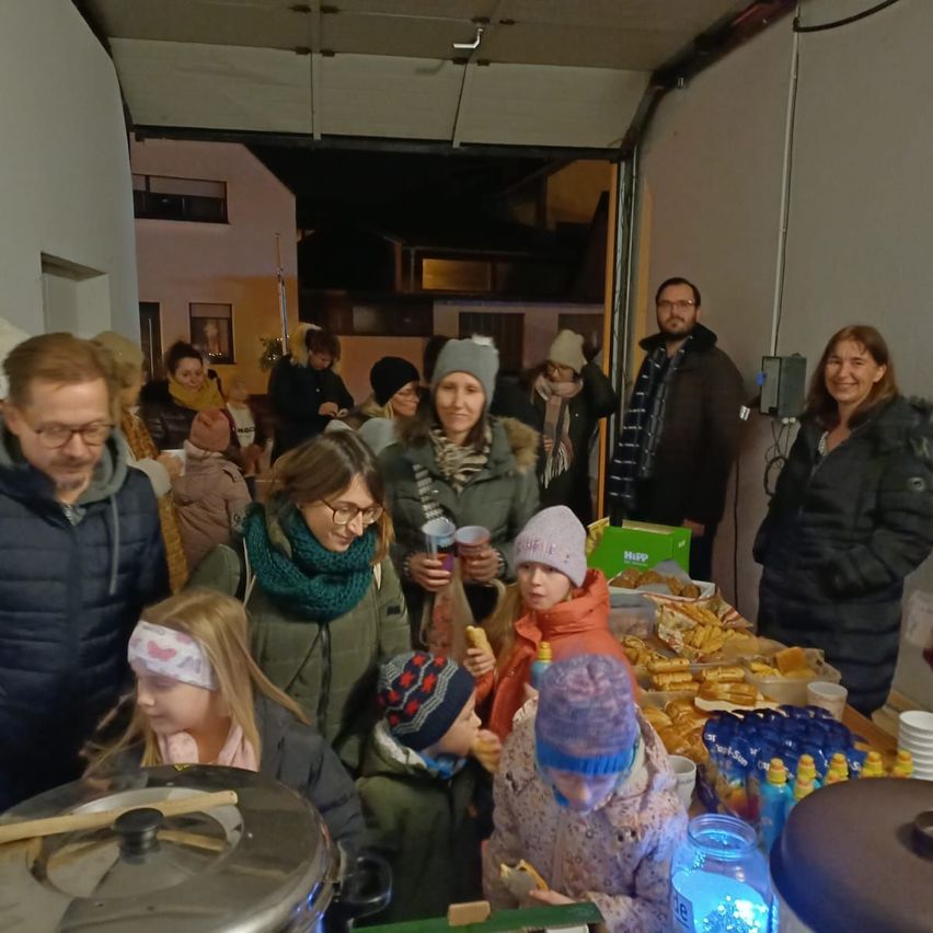 Eine Gruppe von Menschen versammelt sich in einem Raum, einige stehen um einen Tisch mit Essen, andere halten Getränke. Der Raum hat eine Deckenlampe und eine Wandsteckdose.