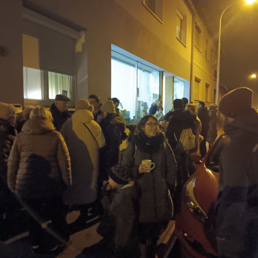 Eine Gruppe von Menschen steht nachts vor einem Gebäude, einige tragen Mützen und halten Tassen, ein rotes Auto ist vor ihnen geparkt.