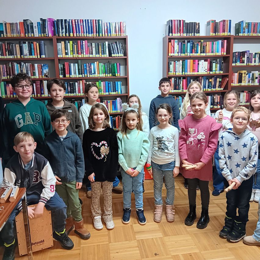 Eine Gruppe junger Kinder steht in einer Bibliothek und lächelt für ein Foto. Einige halten Xylophon, und dahinter sind Bücherregale voller Bücher.