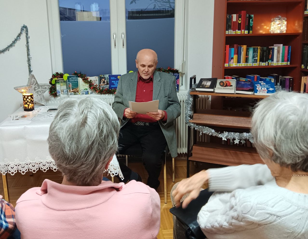 Ein älterer Mann liest ein Stück Papier, während er auf einem Stuhl in einem Raum mit Bücherregalen und dekorativen Gegenständen sitzt. Drei ältere Personen sitzen vor ihm und lauschen.