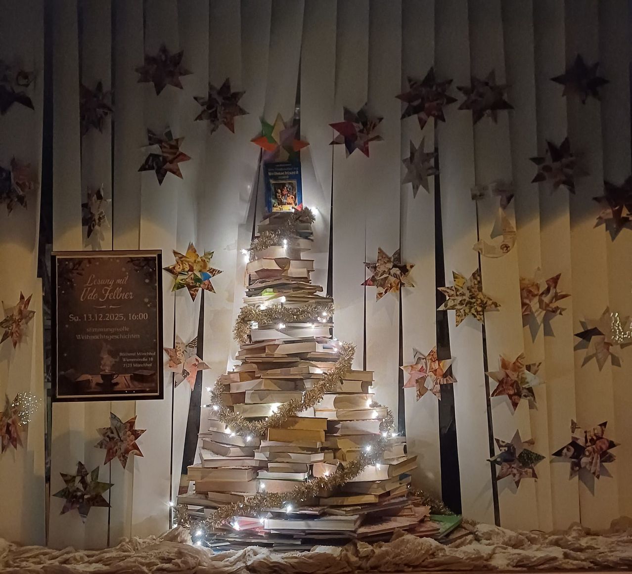 Ein Weihnachtsbaum aus Büchern steht vor einem Vorhang mit Papiersternen. Ein Schild links sagt 'Lesung mit Ude Felner' mit Datum und Uhrzeit.