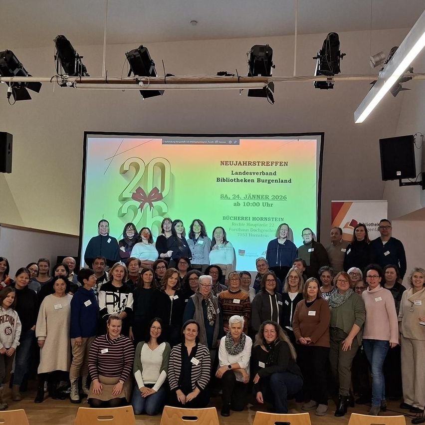 Ein Gruppenfoto von Personen in formeller Kleidung bei einer Neujahrsveranstaltung. Ein Projektionsbildschirm im Hintergrund zeigt ein farbenfrohes Grafik mit dem Namen und Datum der Veranstaltung.