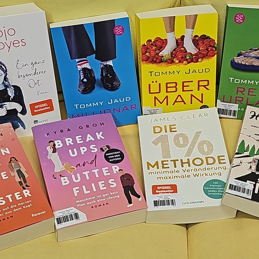 Eine Sammlung von Büchern wird gezeigt, darunter 'Break Ups and Butterflies', 'Die 1% Methode', 'Millionär', 'Über Man', 'Forever' und 'Jo Boyes'. Die Bücher liegen auf einer gelben Oberfläche.