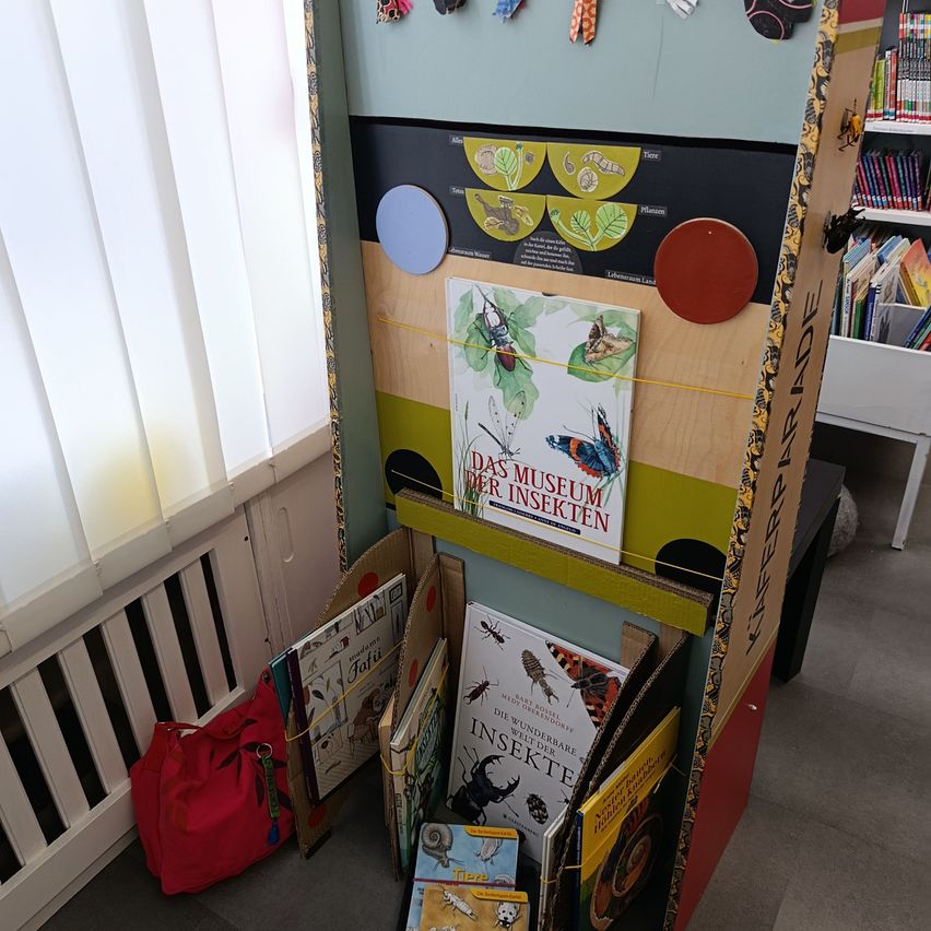 A colorful book display stands in a library, featuring books about insects. A red bag is nearby. The shelves are behind a window with blinds.