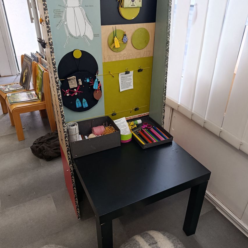 A room with a black table against a wall. A colorful display with various art supplies and a poster of insects is on the table. Wooden chairs and books are placed behind the table.