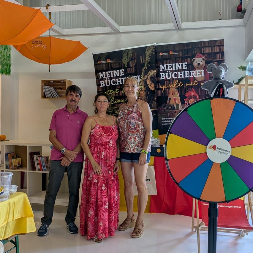 Drei Personen stehen vor einem bunten Rad und einem Banner mit der Aufschrift 'Meine Bucherei spielt mit'. Der Mann trägt ein rosa Poloshirt und die beiden Frauen Kleider. Dahinter steht ein Regal mit Büchern und einer Holzkiste.