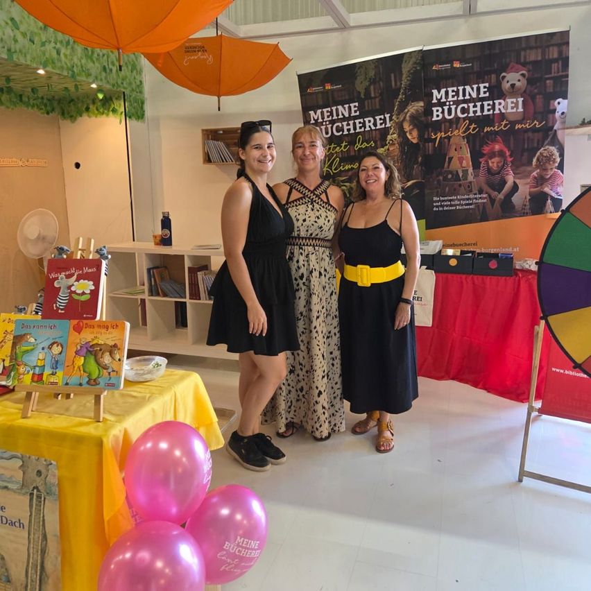 Drei Frauen stehen vor einem Tisch mit Büchern und Luftballons und lächeln für ein Foto. Dahinter hängt ein Banner mit der Aufschrift 'Meine Bucherei' und ein Regal mit Büchern ist sichtbar.