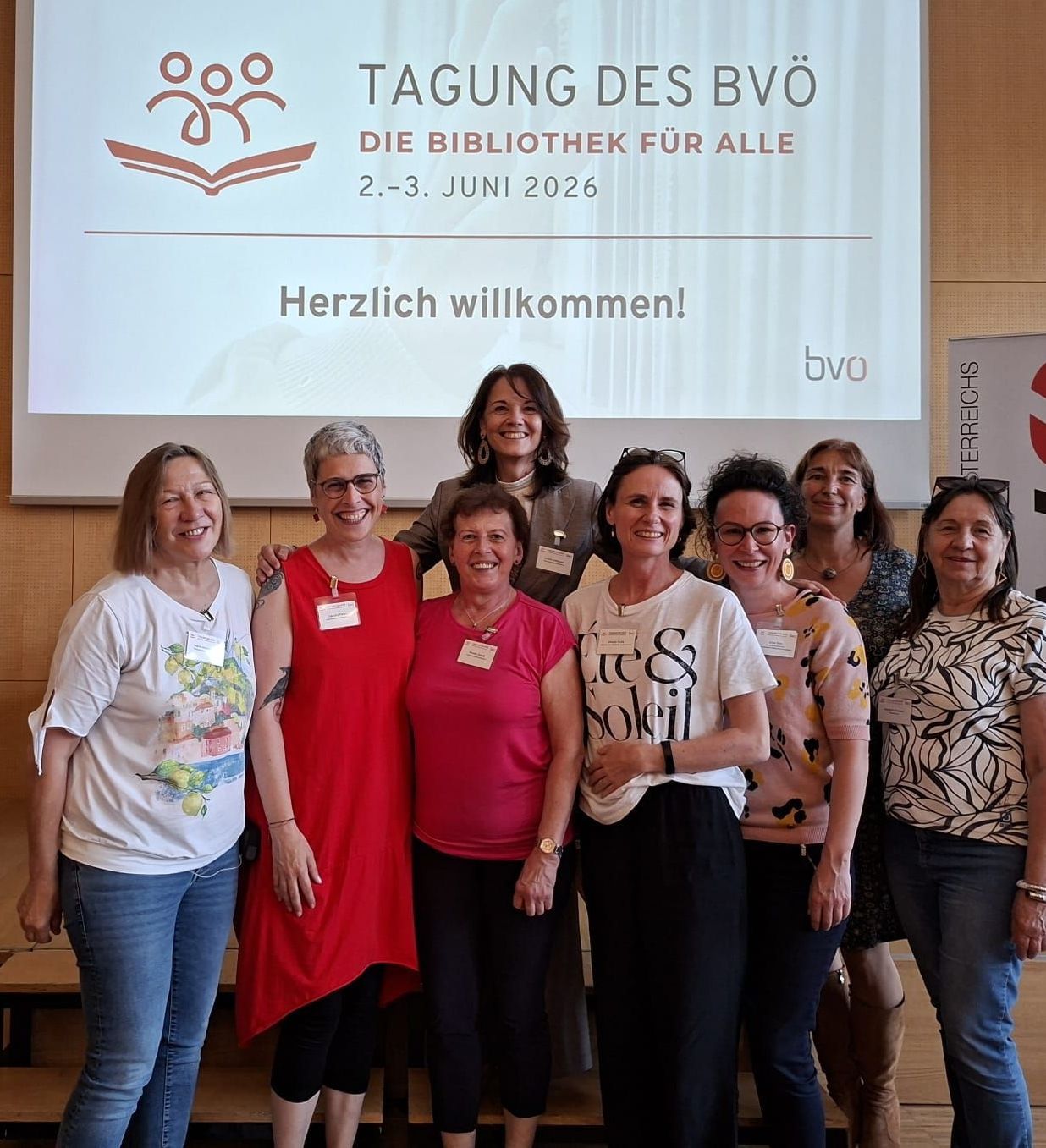 Group photo of women at a library event, smiling, with a projector screen in the background showing an invitation for a library day on June 2-3, 2026.