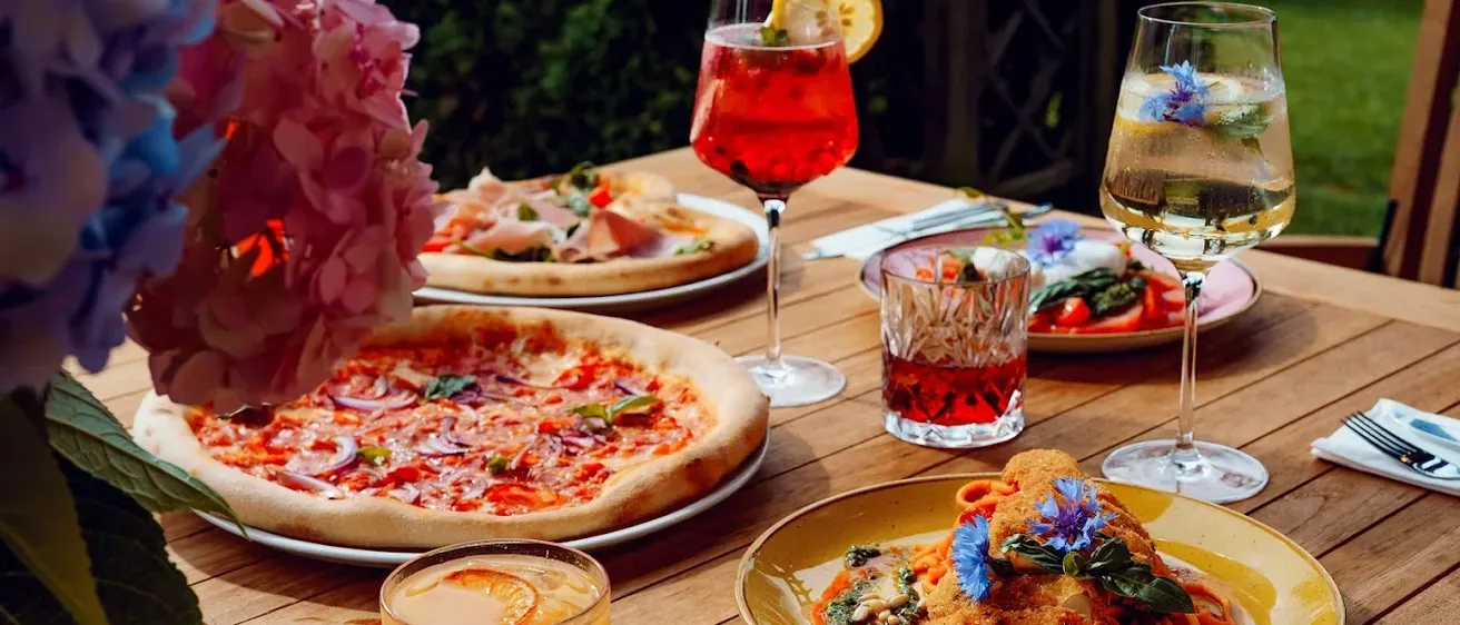 Ein Holztisch ist mit verschiedenen Gerichten und Getränken besetzt, darunter Pizzen, Pasta und Cocktails. Es gibt drei Gläser mit Getränken, einen Teller mit Pizza und einen Teller mit Pasta und Blumen.