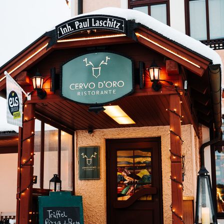 Der Eingang zum CERVO D'ORO RISTORANTE zeigt eine hölzerne Struktur mit einem grünen Schild, dekorativen Lichtern und einem Kreidetafelmenü. Eine Flagge und Lampen befinden sich auf beiden Seiten des Eingangs.