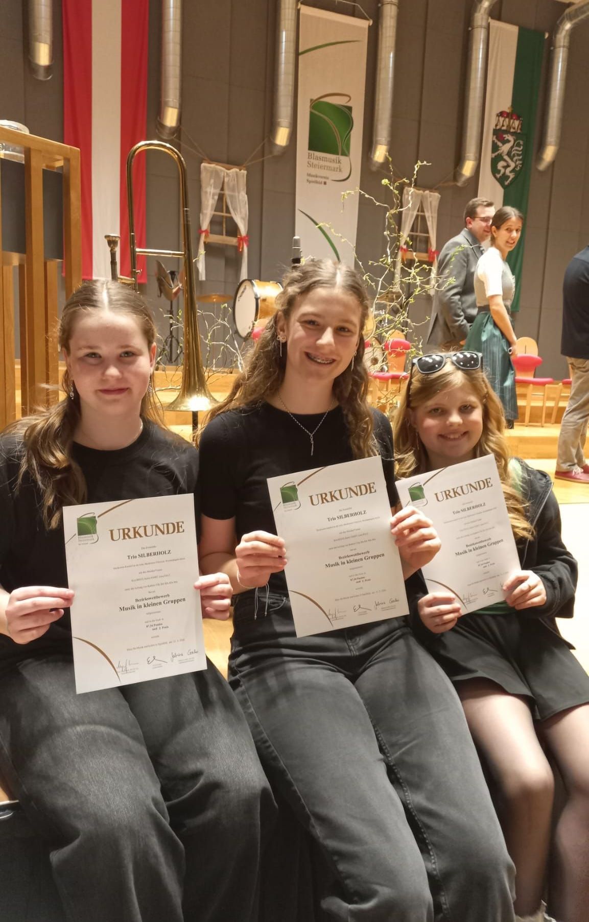 Drei Mädchen sitzen, lächeln und halten Urkunden. Sie tragen Schwarz, eines der Mädchen eine Halskette. Dahinter steht ein Musikinstrumentenaufbau. Zwei Erwachsene stehen im Hintergrund.