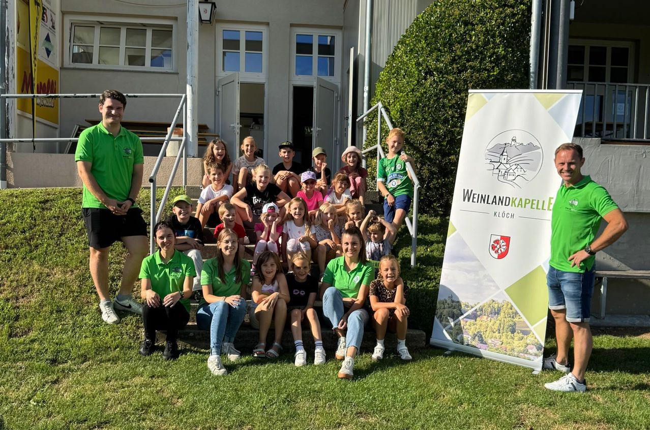 Eine Gruppe von Kindern und Erwachsenen in grünen Shirts posiert für ein Foto vor einem Gebäude mit einem Banner, das 'Weinlandkapelle' steht.