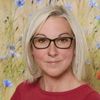 Eine Frau mit blonden Haaren und einer Brille steht in einem Feld mit blauen Blumen und trägt ein rotes T-Shirt.