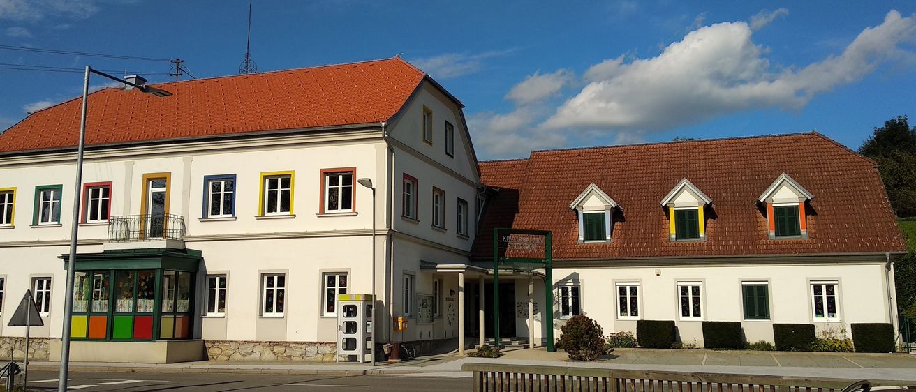 Ein weißes Gebäude mit roten Dächern und gelben Fensterumrandungen. Es befindet sich an einer Straße mit einem Holzzaun davor.