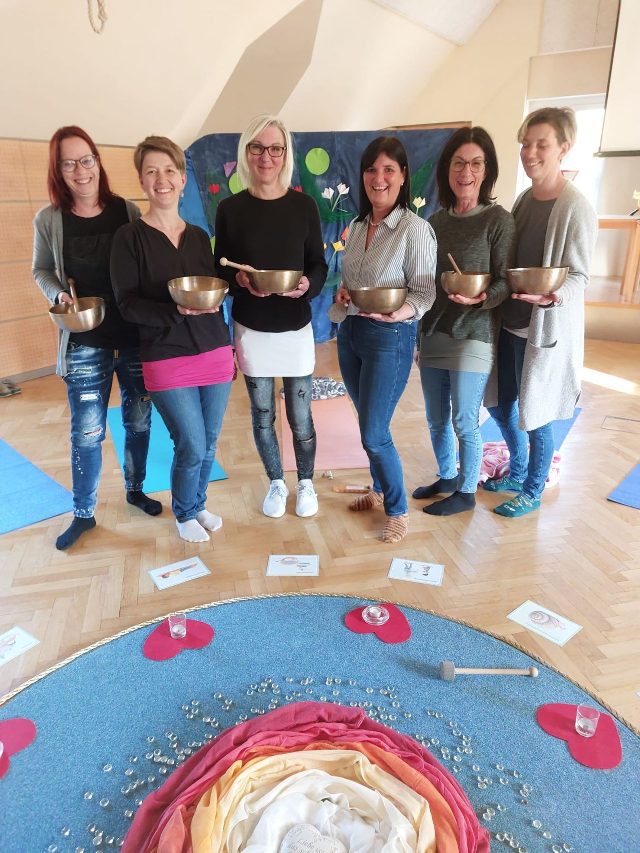 Sechs Frauen in einem Raum mit Holzböden, in Freizeitkleidung und mit Schalen, stehen um einen blauen Kreis mit Glas und einem Holzhammer.