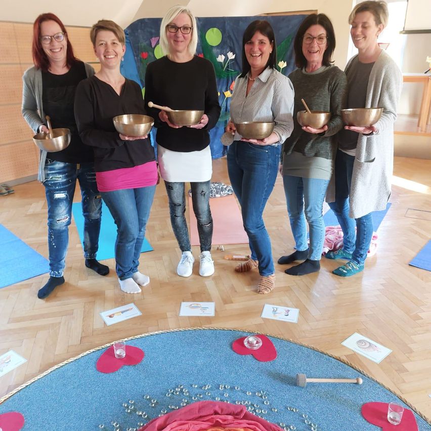 Sechs Frauen in einem Raum mit Holzböden, in Freizeitkleidung und mit Schalen, stehen um einen blauen Kreis mit Glas und einem Holzhammer.