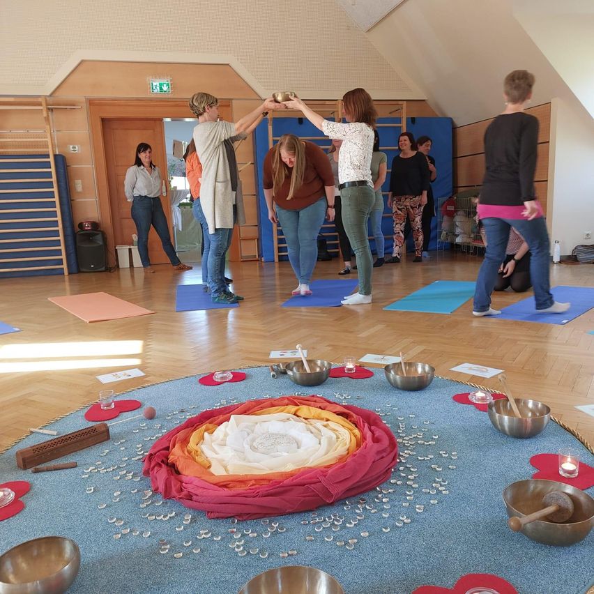 Menschen stehen in einem Kreis in einem Yoga-Studio. Sie halten Schalen und Holzstäbe. In der Mitte des Kreises befindet sich ein roter Stoff mit Kristallen. Yoga-Matten und ein Lautsprecher liegen auf dem Boden.