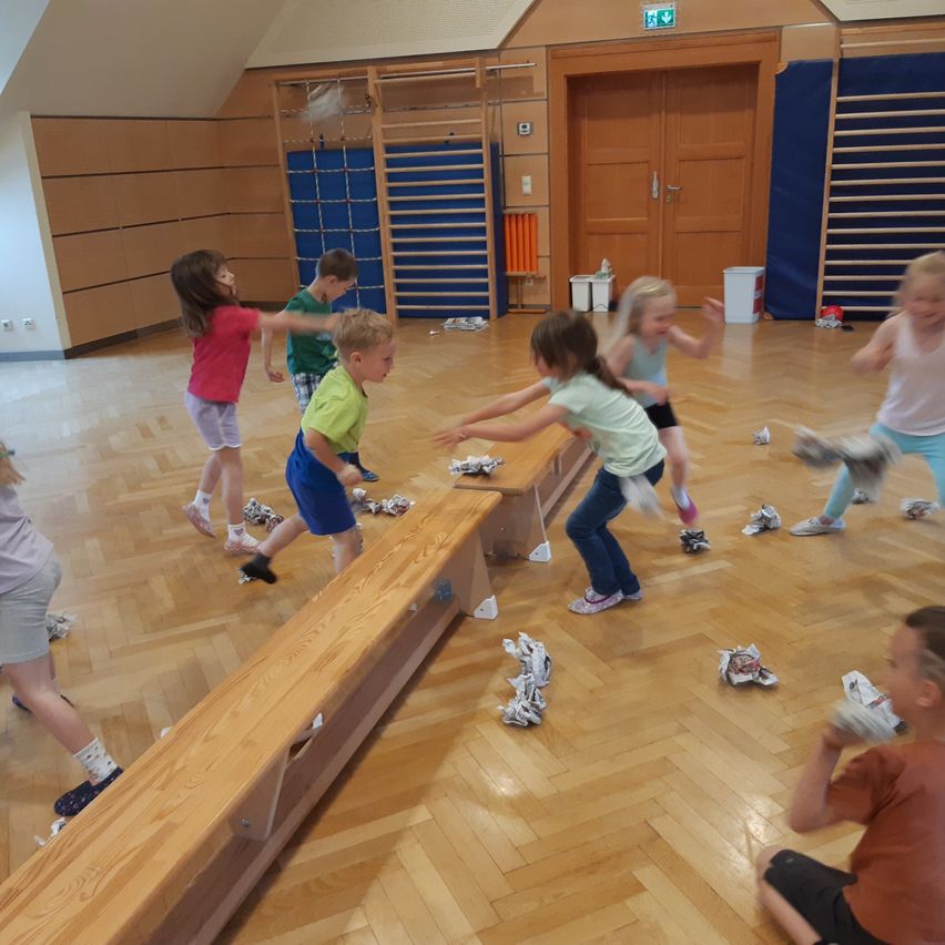 Kinder spielen in einer Turnhalle, einige werfen zerknülltes Papier, während andere lachen und auf einem Holzboden laufen.