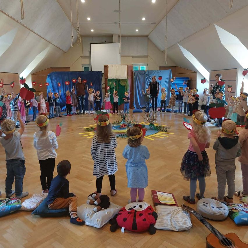 Eine Gruppe von Kindern im Kreis, die Apfelstirnbänder tragen, stehen in einem Raum mit Holzboden und blauem Hintergrund.