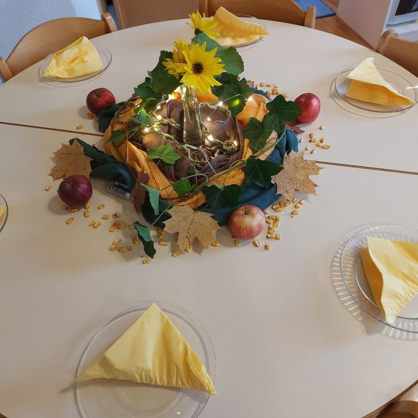 Ein runder Tisch ist mit vier Tellern gedeckt, die jeweils mit einer gefalteten gelben Serviette verziert sind. Im Mittelpunkt steht ein Korb, der in gelbes Tuch gewickelt und mit Sonnenblumen, grünen Blättern und Maiskolben geschmückt ist. Der Tisch ist mit Herbstblättern und Äpfeln dekoriert und schafft eine warme und einladende Atmosphäre.