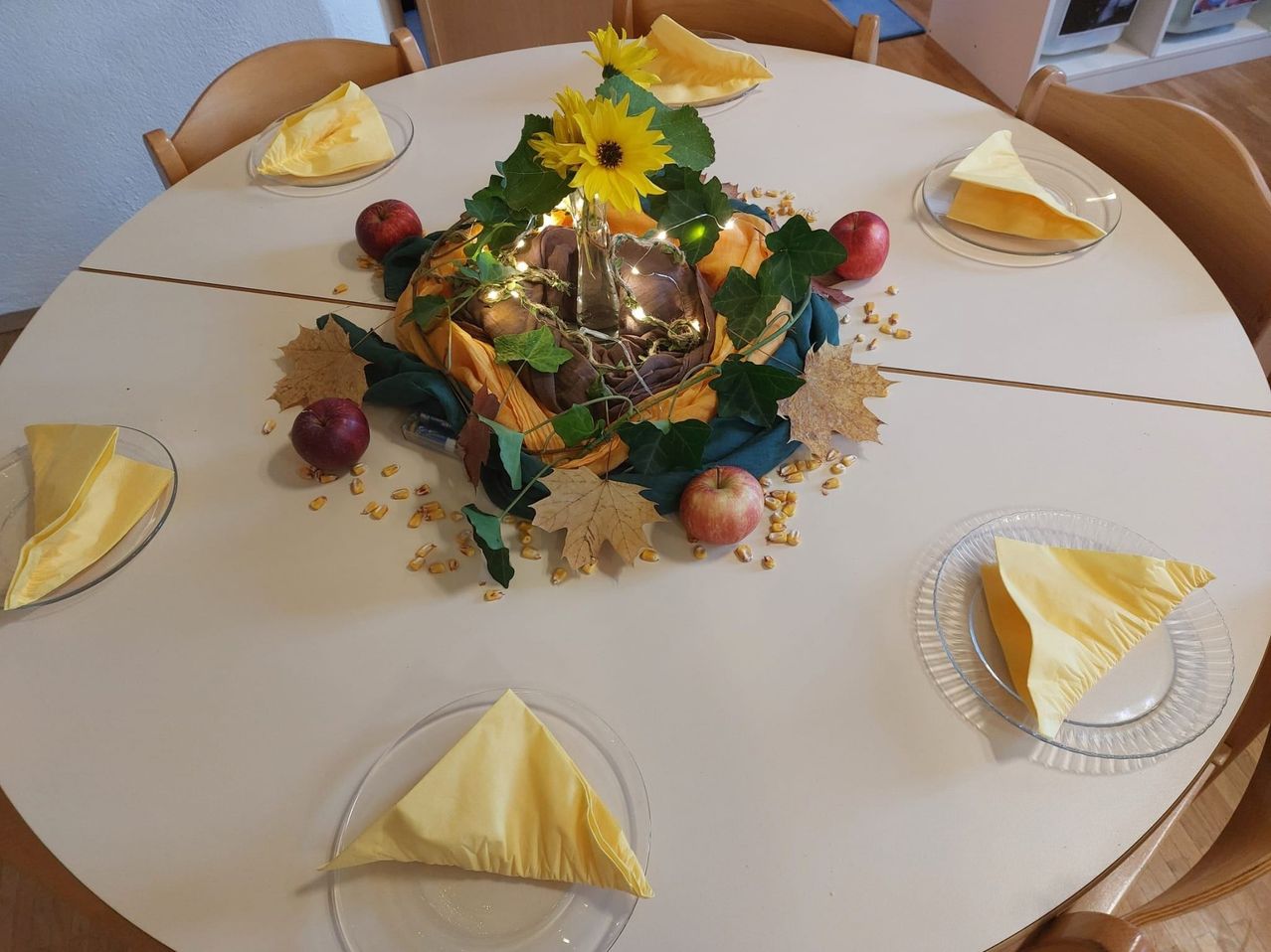 Ein runder Tisch ist mit vier Tellern gedeckt, die jeweils mit einer gefalteten gelben Serviette verziert sind. Im Mittelpunkt steht ein Korb, der in gelbes Tuch gewickelt und mit Sonnenblumen, grünen Blättern und Maiskolben geschmückt ist. Der Tisch ist mit Herbstblättern und Äpfeln dekoriert und schafft eine warme und einladende Atmosphäre.