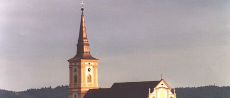 Eine große Kirche mit einem hohen Turm und Uhren steht prominent in einer Stadt, umgeben von Bäumen und Gebäuden.