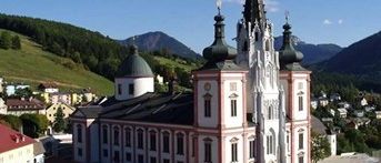 Ein Luftbild einer großen Kirche mit Turm und Kuppeln, umgeben von Gebäuden und Bergen unter einem teilweise bewölkten Himmel.