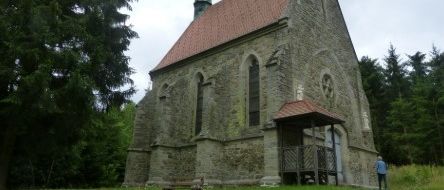 Eine alte Steinkirche mit rotem Dach und einem Turm steht auf einer grasbewachsenen Wiese, umgeben von Bäumen. Eine Person in einem blauen Shirt geht an dem Eingang vorbei.