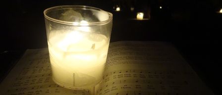Eine Kerze in einem Glas steht auf einem offenen Gesangbuch. Das Buch hat Noten und deutschen Text, beleuchtet durch das Kerzenlicht.