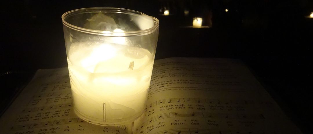 Eine Kerze in einem Glas steht auf einem offenen Gesangbuch. Das Buch hat Noten und deutschen Text, beleuchtet durch das Kerzenlicht.