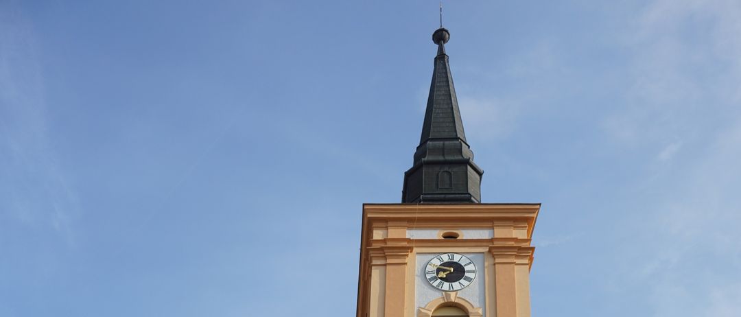 Eine gelbe Kirche mit einem hohen Turm und einer Uhr, umgeben von Bäumen unter einem blauen Himmel.