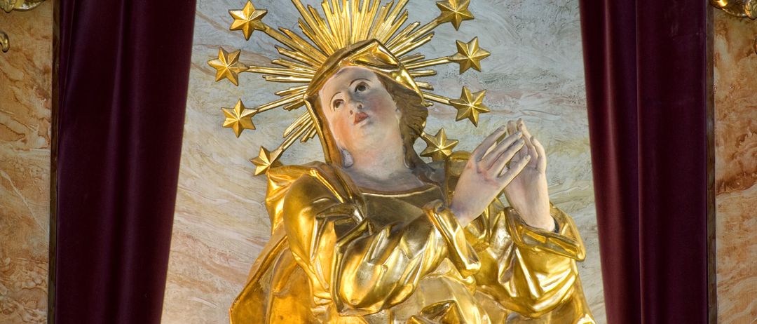 Eine goldene Statue einer Frau mit einem Heiligenschein, der mit Sternen geschmückt ist, trägt ein goldenes Kleid und betet mit gefalteten Händen.