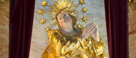 Eine goldene Statue einer Frau mit einem Heiligenschein, der mit Sternen geschmückt ist, trägt ein goldenes Kleid und betet mit gefalteten Händen.