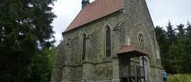 Eine alte Steinkirche mit rotem Dach und einem Turm steht in einem grasigen Feld, umgeben von Bäumen. Eine Person geht in der Nähe des Eingangs.