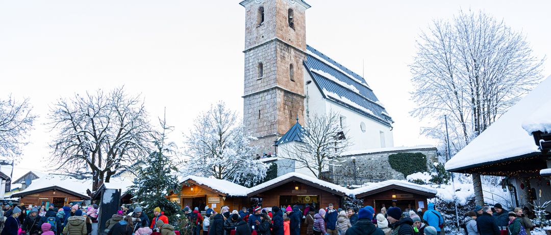 Eine Menschenmenge versammelt sich um eine Kirche im Schnee. Die Kirche hat einen hohen Turm und eine Glocke. Es ist eine Winterszene mit Schnee auf den Bäumen und dem Boden.