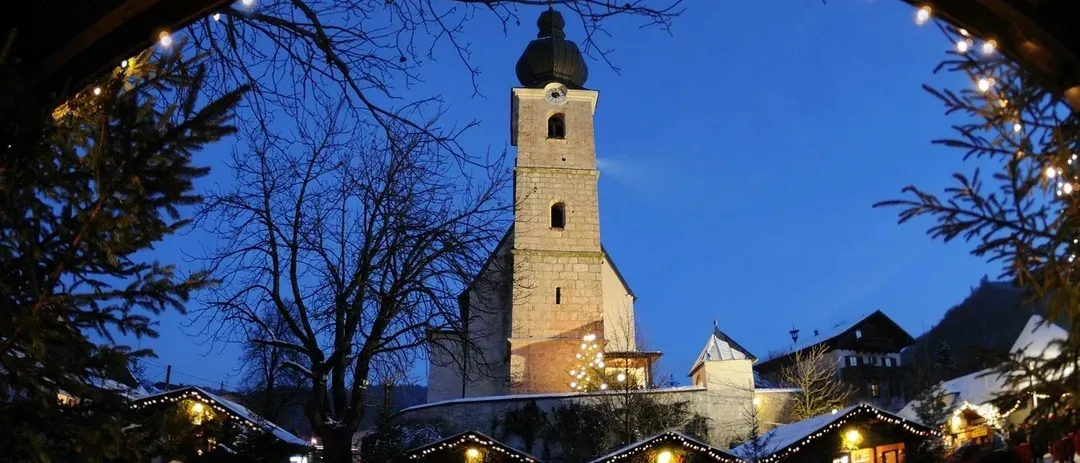 Ein Weihnachtsmarkt vor einer Kirche mit einem hohen Glockenturm ist bei Nacht beleuchtet. Menschen gehen herum, und es gibt Stände mit Lichtern.