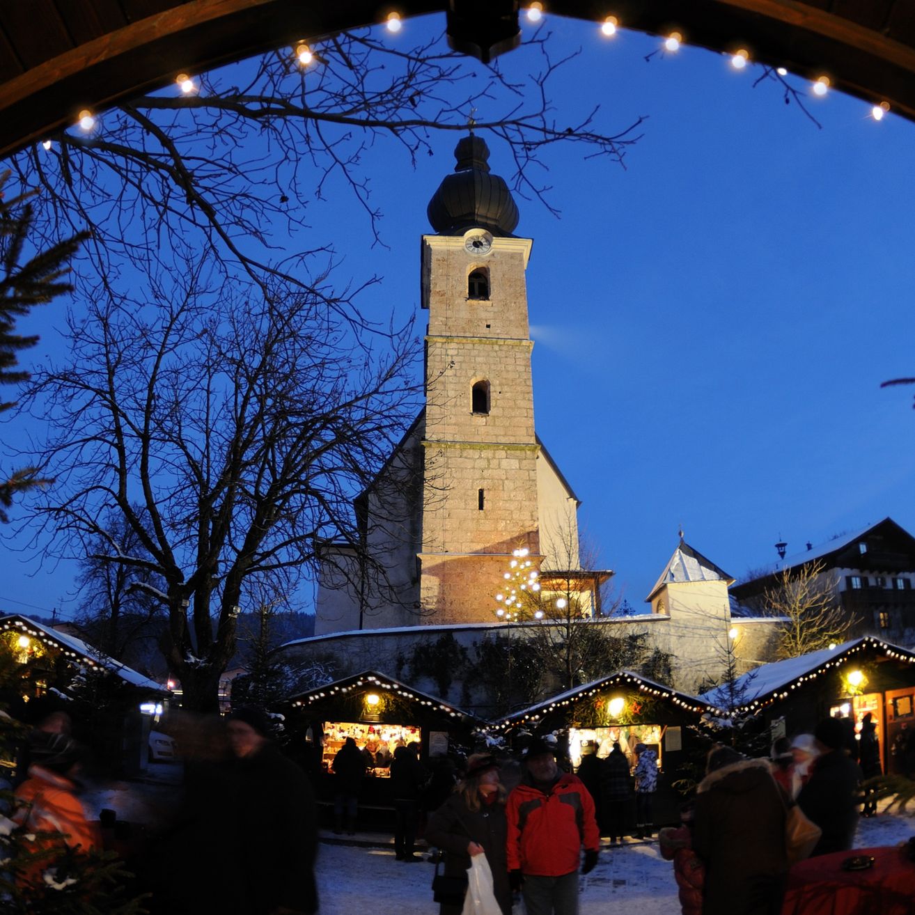 Bei Dämmerung versammeln sich Menschen auf einem festlichen Weihnachtsmarkt vor einer historischen Kirche, deren Turm mit einem Weihnachtsbaum und funkelnden Lichtern geschmückt ist.