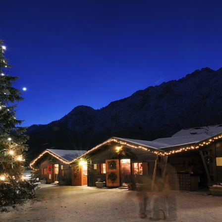 Ein Weihnachtsbaum ist vor einer gemütlichen Hütte beleuchtet, mit Weihnachtsdekorationen und Lichtern auf dem Dach. Verschneite Berge sind im Hintergrund zu sehen, und eine Person steht im Vordergrund.