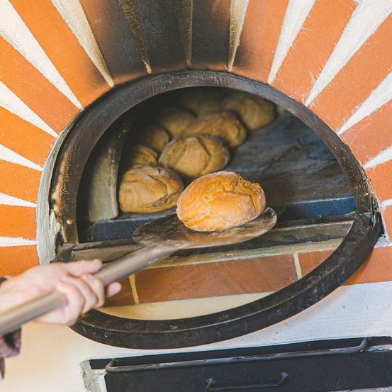 Eine Person benutzt einen Spatel, um ein Brot aus einem Backofen zu nehmen. Der Ofen ist mit anderen Broten gefüllt und hat ein orange-weiß gestreiftes Äußeres.