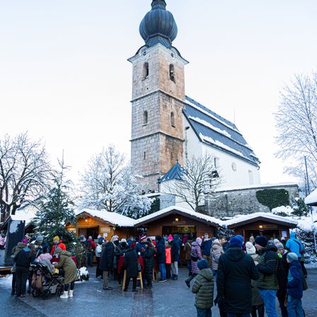 Eine Menschenmenge versammelt sich auf einem Weihnachtsmarkt vor einer schneebedeckten Kirche.