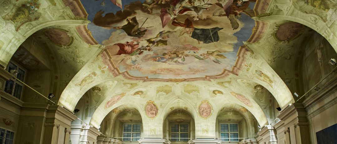 The interior of a church features a detailed ceiling fresco depicting various figures in the sky. Below, three arched windows illuminate the nave. The walls are adorned with decorative elements, and an ornate organ is prominently displayed.