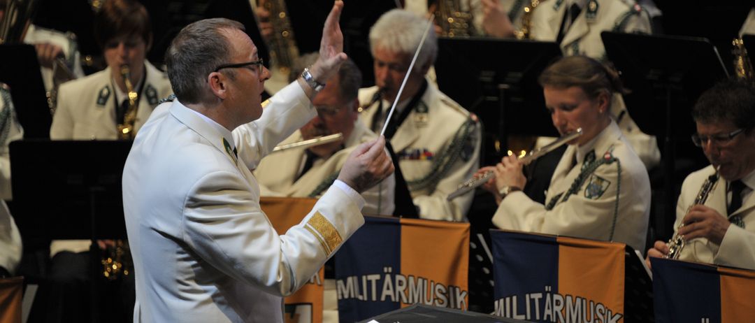A military band performs on stage, led by a conductor in a white uniform, holding a baton. Behind him, band members play various instruments, including trumpets and flutes. Banners with military music insignia are visible on stage.