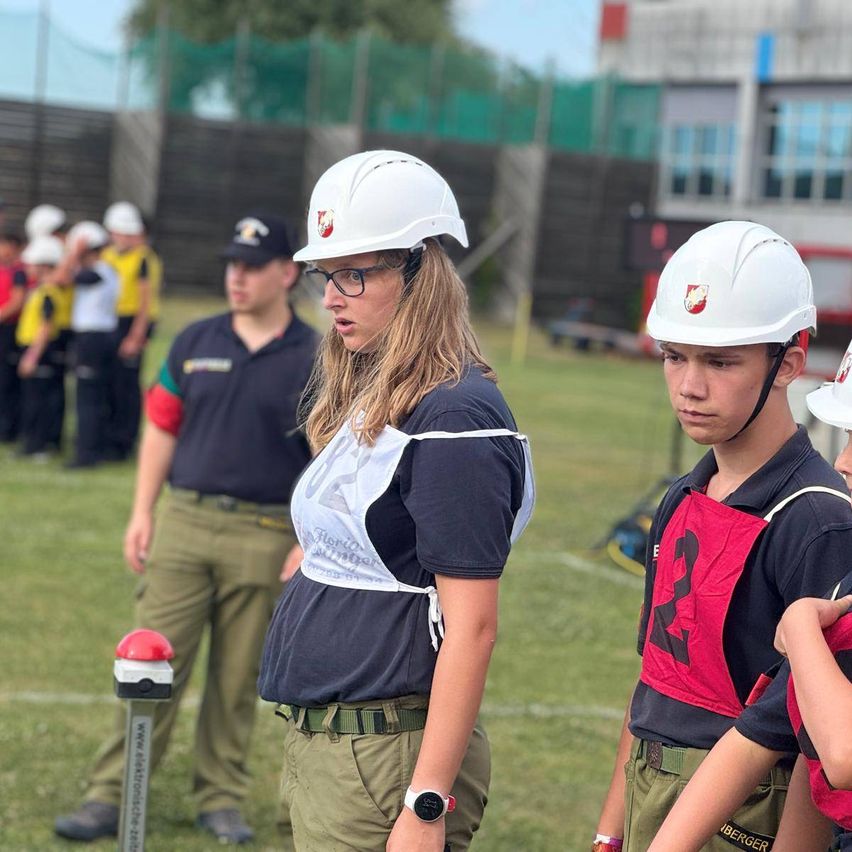 Eine Gruppe von Menschen in Schutzhelmen und Uniformen steht auf einem Rasenfeld. Eine Frau schaut etwas vor sich an. Ein Junge steht neben ihr. Dahinter stehen Menschen in einer Reihe.