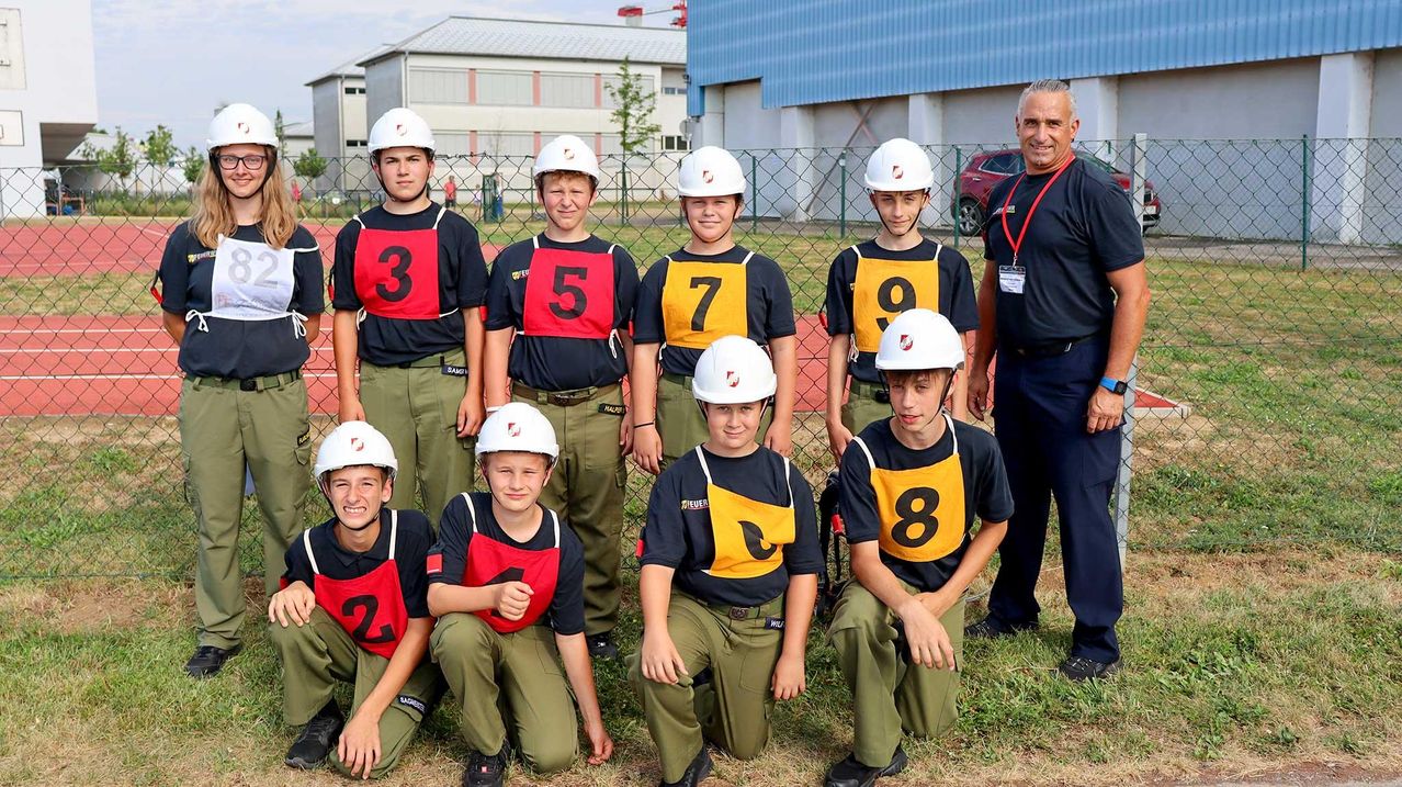 Eine Gruppe junger Jungen in nummerierten Uniformen und Helmen posiert für ein Foto auf einem Feld. Ein Mann steht hinter ihnen.