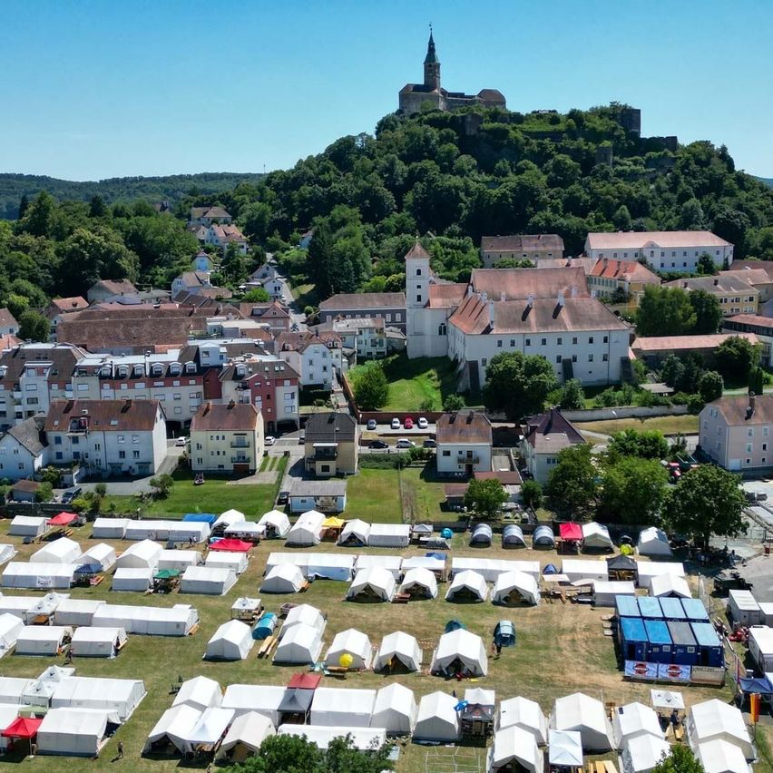 Luftaufnahme einer Stadt mit vielen Zelten auf einem Feld, umgeben von Bäumen und Häusern. Ein Schloss steht auf einem Hügel in der Ferne.